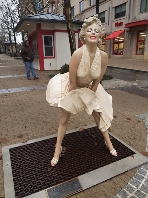 A statue of Marilyn Monroe in a flowing beige dress, playfully holding it up, set in an outdoor plaza.