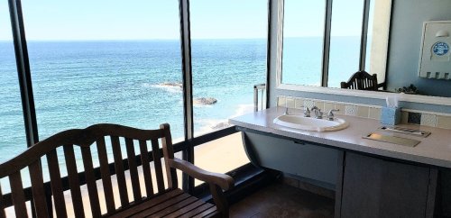 A bright bathroom with a sink and a wooden bench, featuring large windows overlooking the ocean.