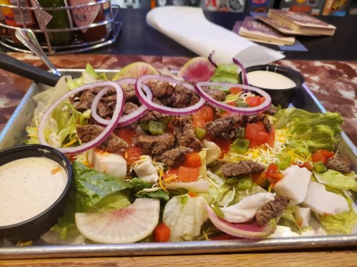 A colorful salad with lettuce, tomatoes, steak, cheese, and radish, served with two dressing cups on a metal tray.