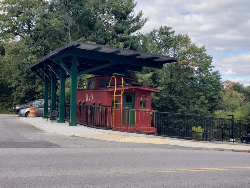 A red caboose on a platform with a roof, surrounded by trees and parked cars, under a cloudy sky.