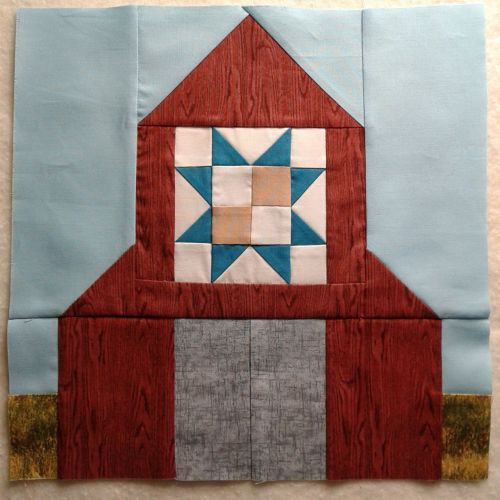 A quilt block featuring a barn design with a star pattern, set against a light blue background and grassy edges.