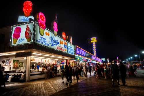 Brightly lit boardwalk ice cream stand at night, bustling with people enjoying treats and vibrant neon signs.