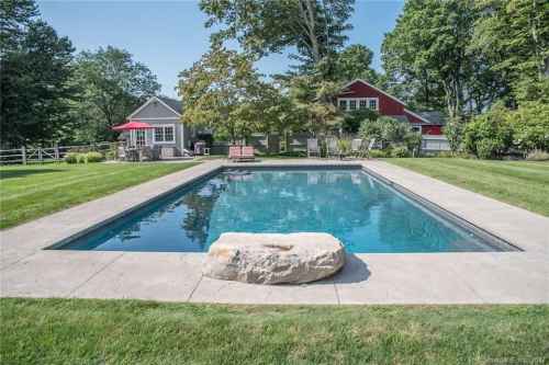 A serene backyard with a rectangular pool, surrounded by green grass and trees, featuring a rock and nearby seating areas.