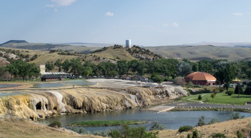 A scenic view of a landscape featuring colorful rock formations, a river, and distant hills under a clear blue sky.