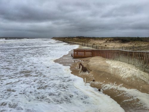 Waves crash onto a sandy beach with a wooden fence, under a cloudy sky. Coastal erosion is visible along the shoreline.