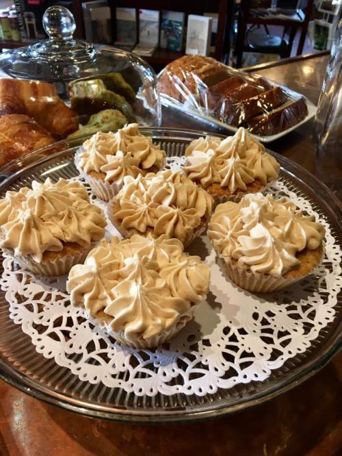 A platter of six cupcakes with swirled caramel frosting, surrounded by baked goods in a cozy café setting.