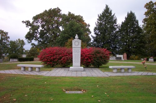A memorial surrounded by vibrant red bushes and benches, set in a grassy area with trees in the background.
