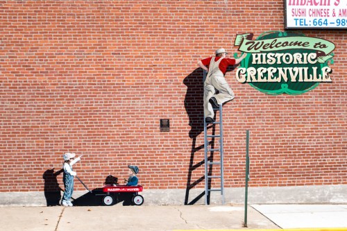 A mural depicting a man on a ladder painting a sign, with two children in a wagon below, against a brick wall.