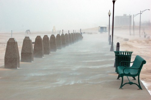 A foggy boardwalk with stone barriers and a green bench, surrounded by blowing sand and a distant building.