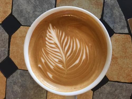 A close-up of a latte with intricate leaf latte art in a white cup on a patterned tile surface.