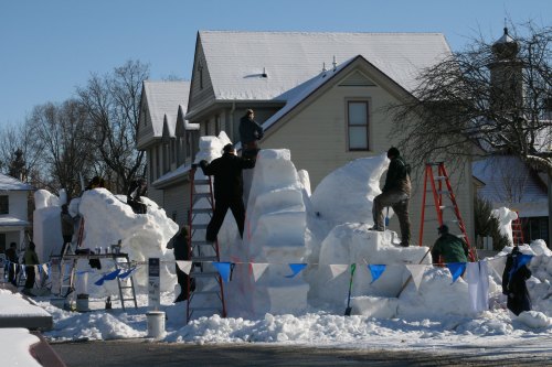 People building large snow sculptures in a snowy neighborhood, with ladders and colorful flags around the site.