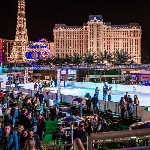 A lively outdoor ice skating rink at night, with a city skyline and festive lights in the background.