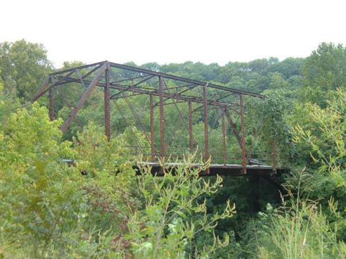 An old, rusted bridge covered in greenery, surrounded by dense trees and foliage.