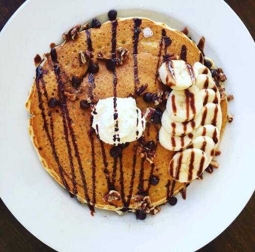 A plate of pancakes topped with banana slices, chocolate drizzle, whipped cream, and chopped nuts.