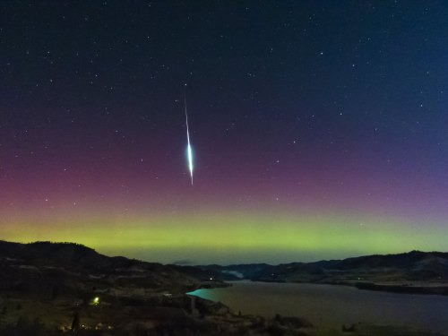 A stunning night sky with a bright meteor streaking above a colorful aurora over a calm lake and hills.