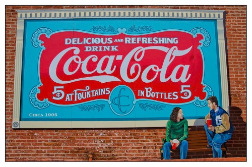 Vintage Coca-Cola mural featuring two people sitting and enjoying drinks, with a retro design and text from 1905.