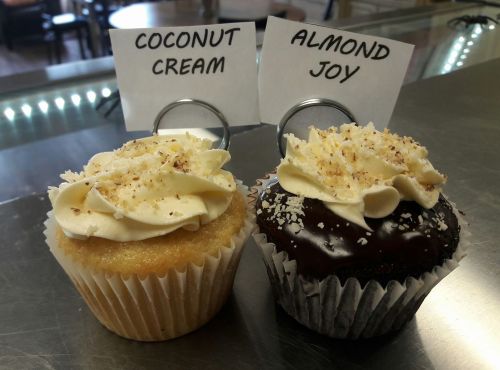 Two cupcakes on a counter: one coconut cream topped with frosting, the other chocolate almond joy with a similar frosting.