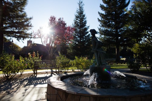 A sunlit park scene featuring a fountain with a statue, surrounded by trees and blooming flowers.