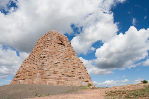 A large stone pyramid structure under a blue sky with fluffy clouds, surrounded by a rocky landscape.