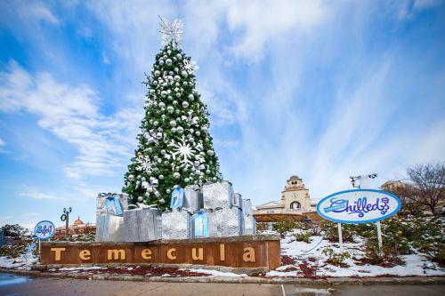 A large decorated Christmas tree with presents at the base, in front of a sign for Temecula, under a blue sky.