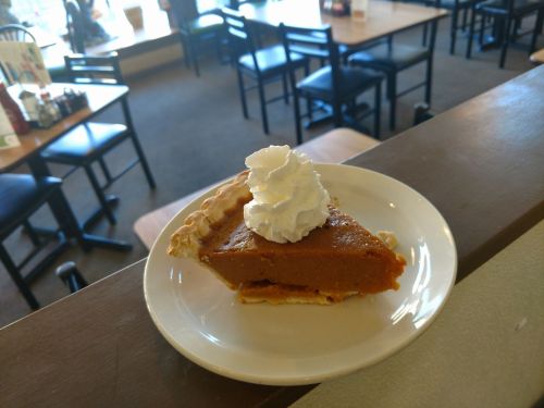 A slice of pumpkin pie topped with whipped cream on a white plate, with a restaurant interior in the background.