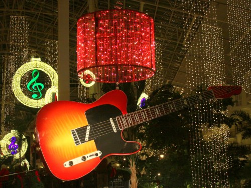 A large, vibrant orange guitar hangs amidst festive lights and decorations in a holiday setting.