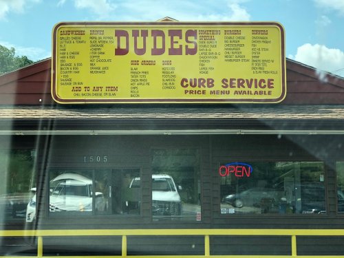 Sign for "Dudes" featuring a menu with sandwiches, drinks, and sides, with an "Open" neon sign visible.