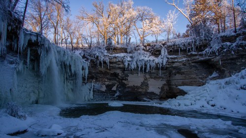 A frozen waterfall surrounded by icy cliffs and snow-covered trees under a clear blue sky.