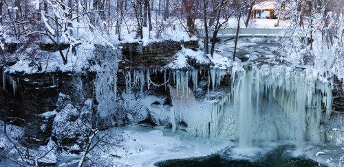 A winter scene of a frozen waterfall surrounded by snow-covered trees and icicles, with a bridge in the background.