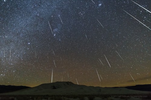 A starry night sky filled with meteor trails above a dark, silhouetted landscape.