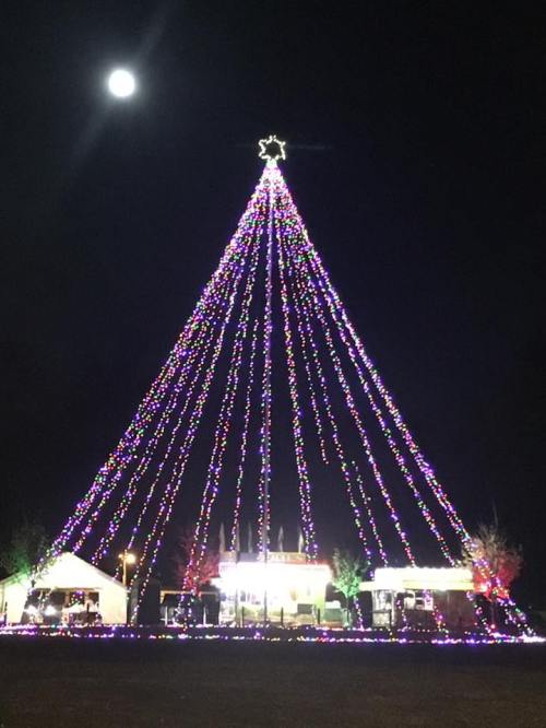 A tall, illuminated Christmas tree made of colorful lights, with a star on top, set against a night sky and moon.