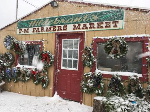 A snowy scene of Hildebrandt's F-M Farmers Market, decorated with wreaths and a welcoming "Open" sign.