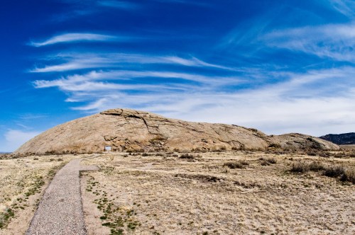A large rock formation under a blue sky with wispy clouds, surrounded by dry grass and a gravel path.