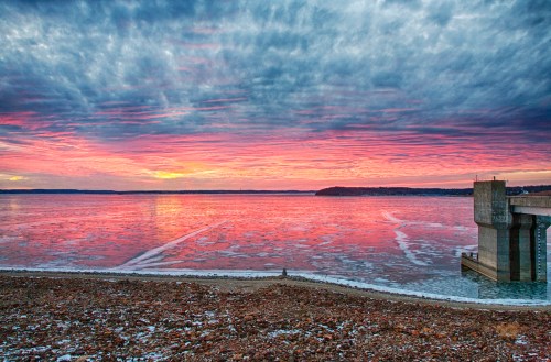 A vibrant sunset over a frozen lake, with colorful clouds reflecting on the icy water and a concrete pier in the foreground.