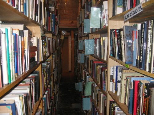 A narrow aisle between tall bookshelves filled with various books, leading to a dimly lit area at the end.
