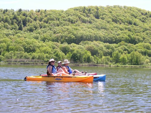 Three people in kayaks on a calm lake, surrounded by lush green trees and hills under a clear blue sky.