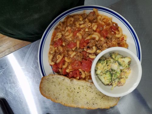 A plate of macaroni with meat sauce and tomatoes, accompanied by a side of broccoli salad and a slice of garlic bread.