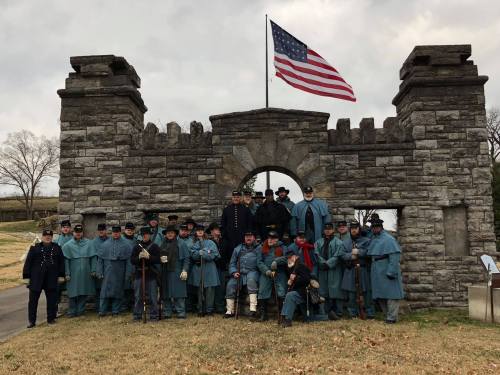 A group of historical reenactors in Civil War uniforms poses in front of a stone archway with an American flag.