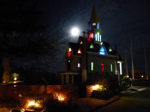 A brightly lit, colorful church at night with a full moon in the background and illuminated pathways.