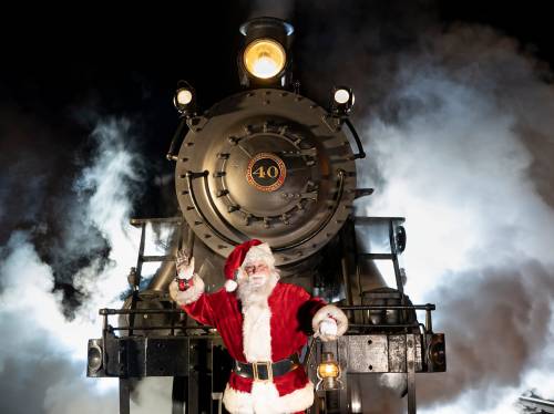 Santa Claus waves in front of a vintage steam train, surrounded by billowing smoke and festive lights.