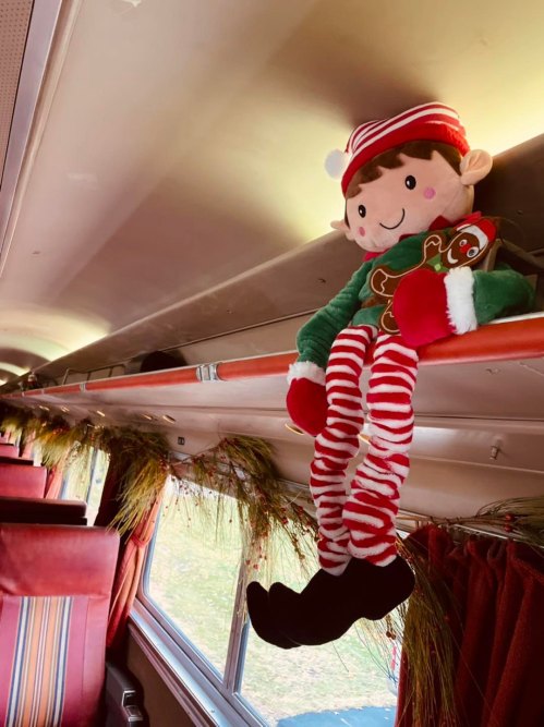 A plush elf doll holding a gingerbread cookie sits on a train's overhead rack, decorated with festive greenery.