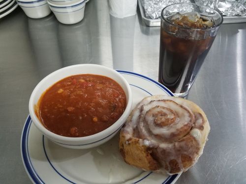 A bowl of chili, a cinnamon roll, and a glass of iced drink on a plate.