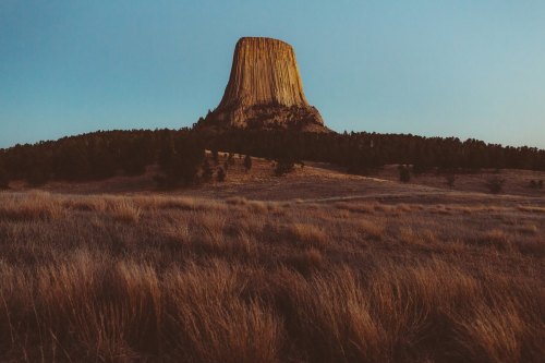 A towering rock formation rises above a grassy landscape under a clear blue sky at sunset.