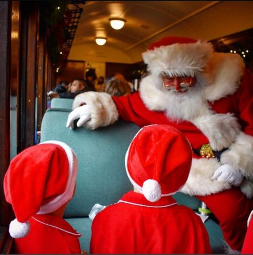 Santa Claus interacts with two children in red outfits on a festive train, surrounded by holiday decorations.