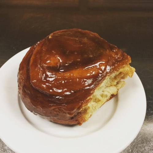 A large, glossy cinnamon roll on a white plate, showcasing its sticky glaze and fluffy texture.