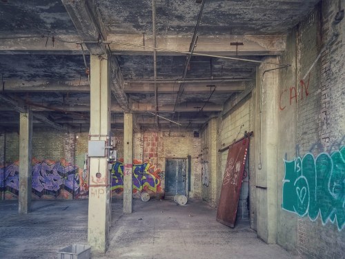 Abandoned industrial space with concrete pillars, graffiti on walls, and scattered debris on the floor.