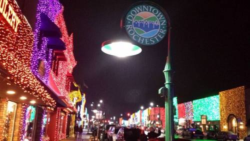 Colorful holiday lights adorn buildings along a street in downtown Rochester at night, with a sign overhead.