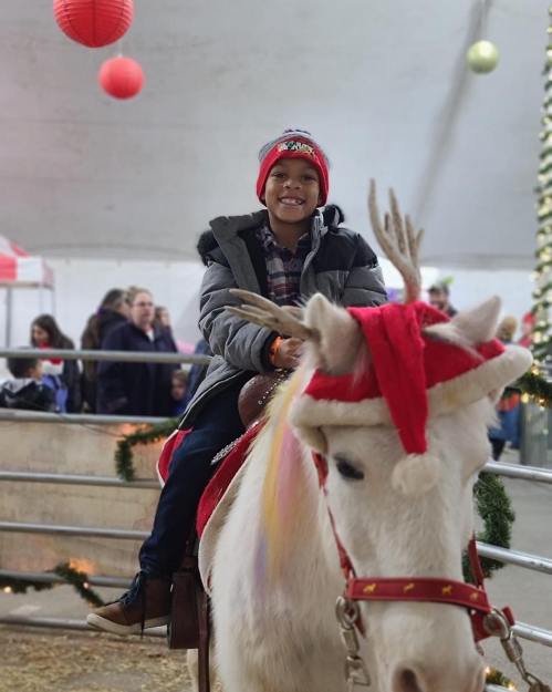 A smiling child in a winter coat rides a white pony adorned with festive decorations at a holiday event.