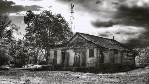 A weathered, abandoned building with a metal roof, surrounded by trees and under a dramatic sky, in black and white.