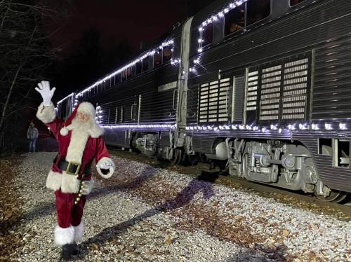 A festive Santa waves near a train adorned with holiday lights, surrounded by a wooded area at night.